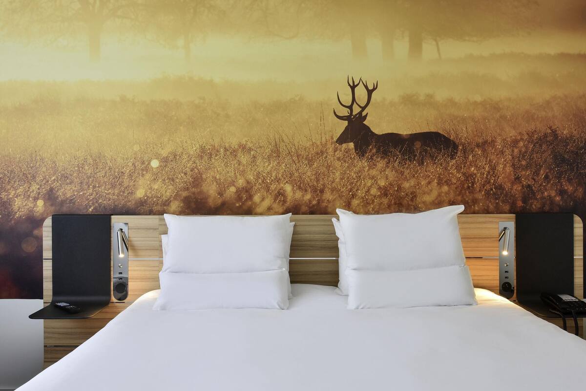 Habitación de hotel con una cama grande, dos almohadas blancas, un cabecero con un teléfono incorporado y un mural en la pared de un ciervo en un campo.