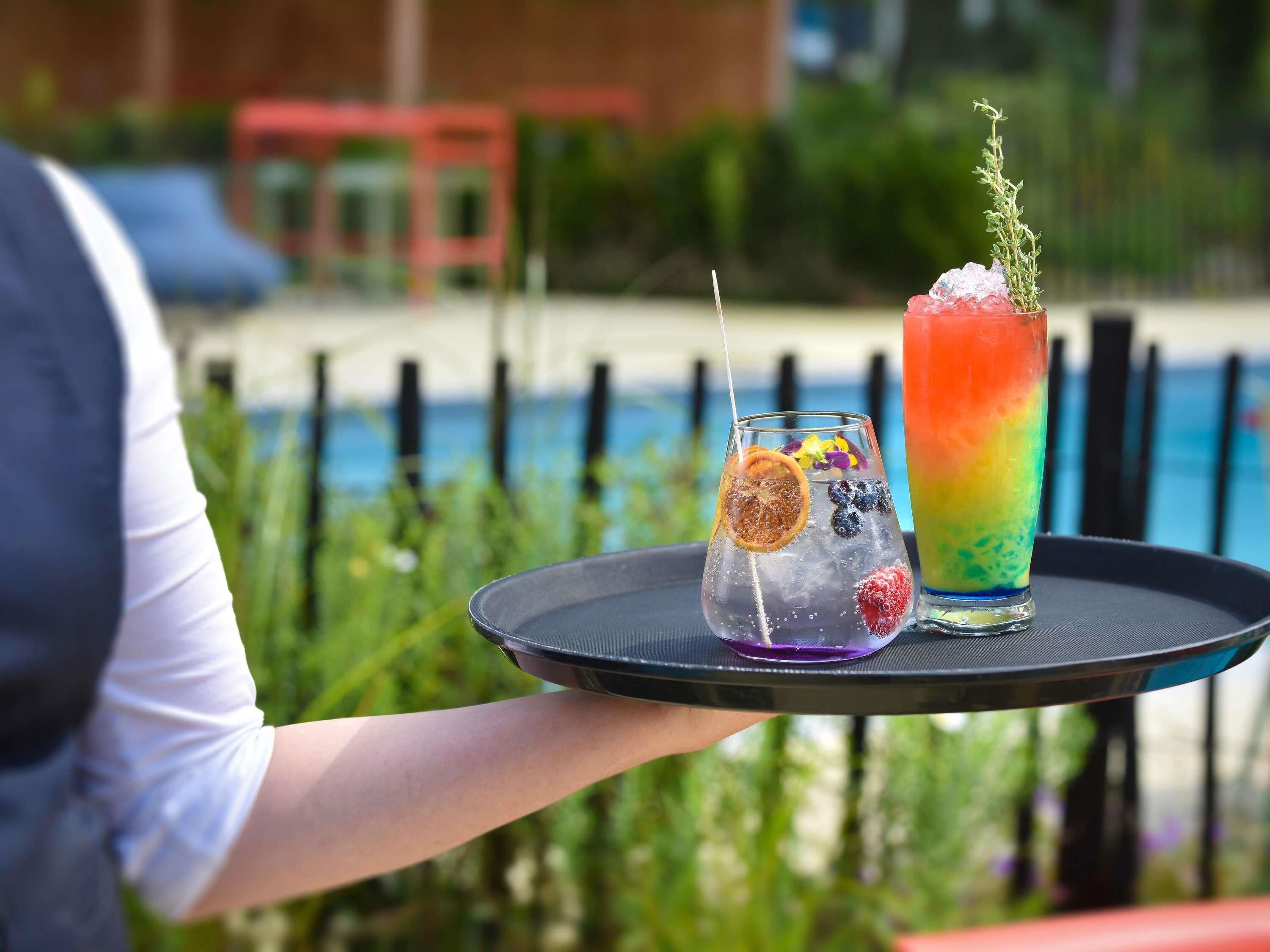 A tray with two drinks, one with a slice of fruit and the other with a colorful layered drink, held by a person.