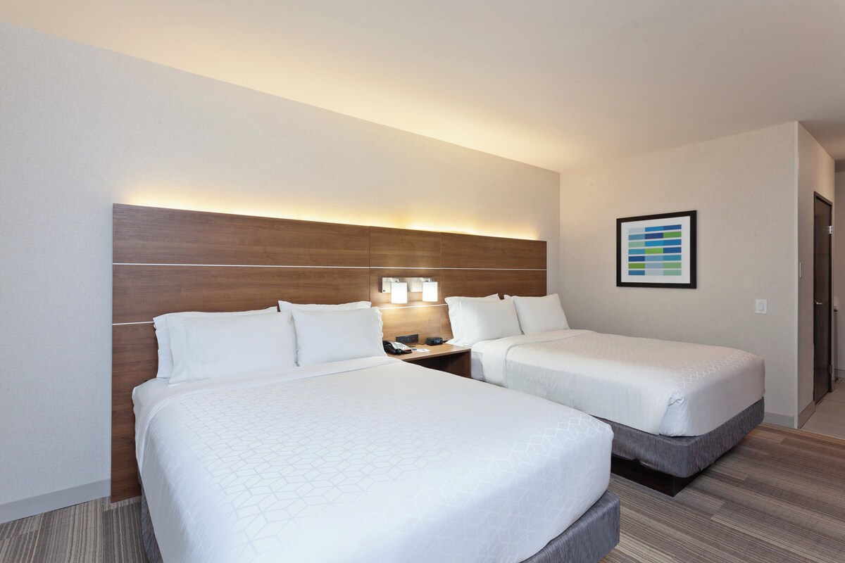 A hotel room with two beds, a nightstand, a wall-mounted lamp, and a framed picture on the wall.