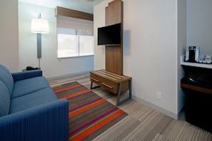 A modern hotel room with a blue sofa, a wooden desk, a flat-screen TV, a coffee machine, and a colorful striped rug. - Holiday Inn Express Los Angeles Airport by IHG (Los Angeles)