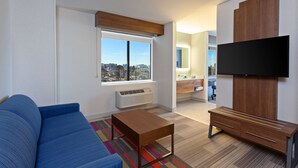 Un salon moderne avec un canapé bleu, une table basse en bois, un téléviseur à écran plat fixé sur un meuble en bois et une grande fenêtre donnant sur la ville.