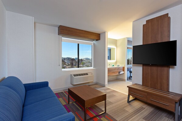 A modern living room with a blue sofa, a wooden coffee table, a flat-screen TV mounted on a wooden cabinet, and a large window with a city view. - Holiday Inn Express Los Angeles Airport by IHG (Los Angeles)