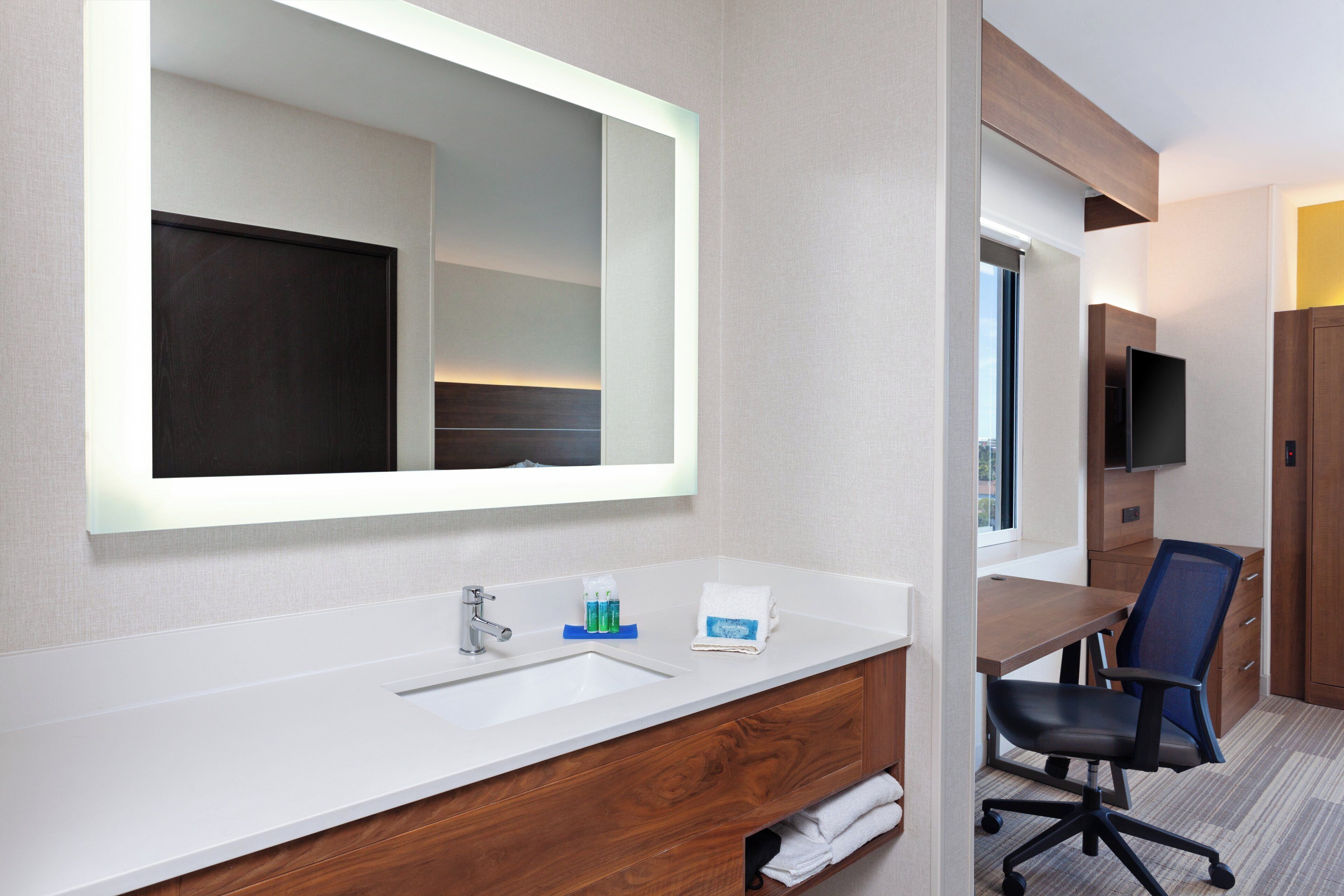 A hotel bathroom with a large mirror, a sink, a vanity, a chair, and a television.