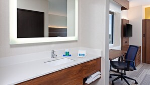 Une salle de bain d’hôtel avec un grand miroir, un lavabo, une commode, une chaise et un téléviseur.