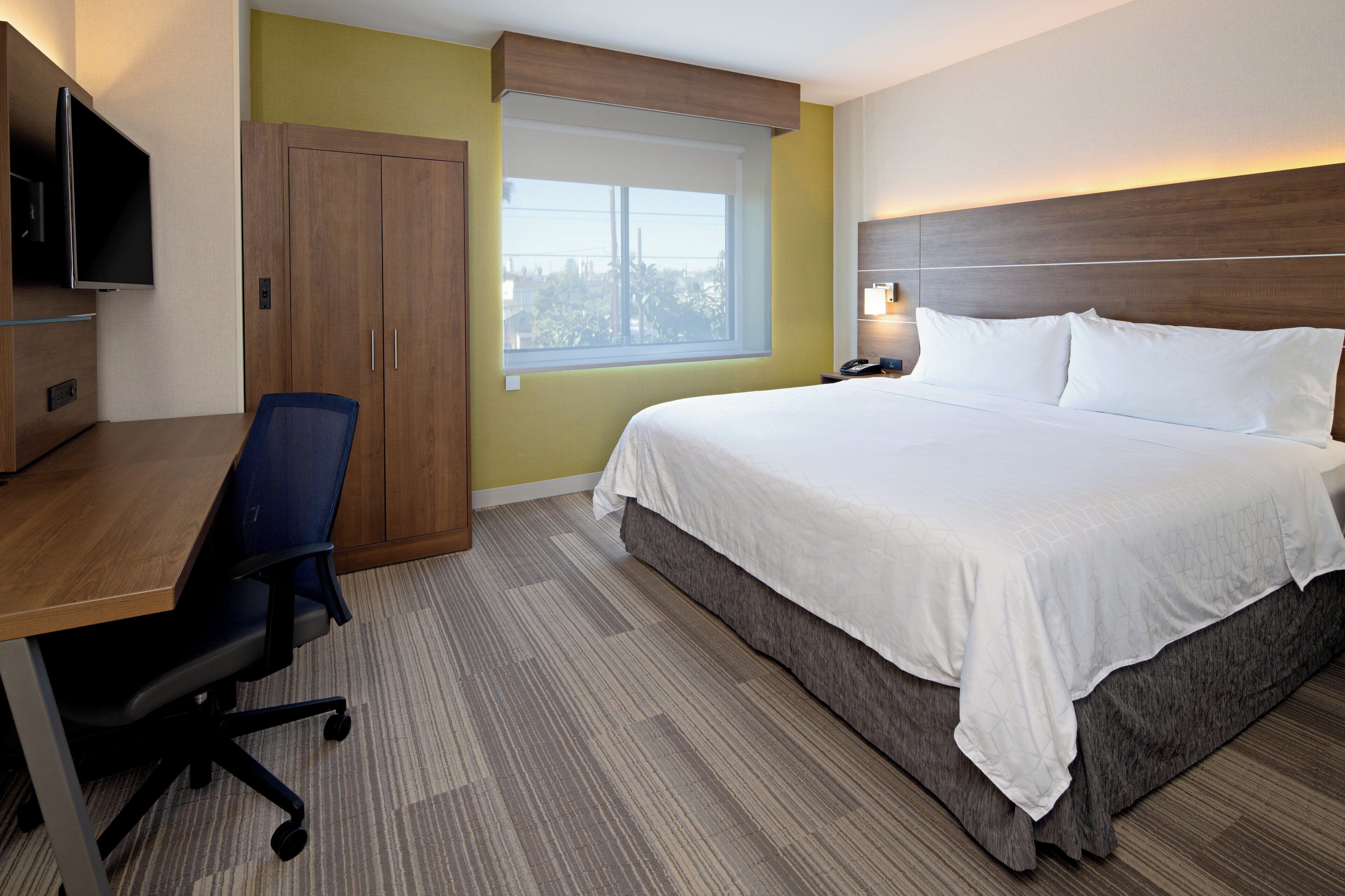 Habitación de hotel con cama, escritorio, silla, televisión, ventana y mesita de noche con lámpara.