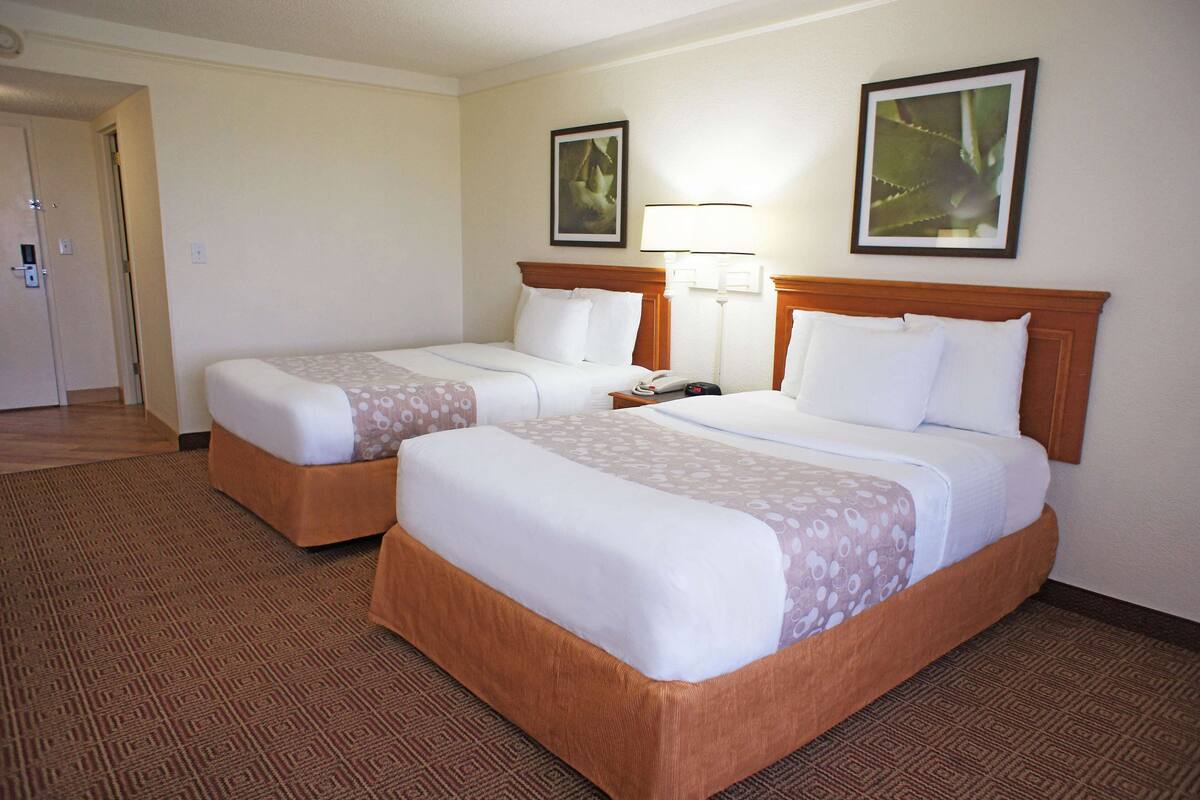 A hotel room with two beds, a wooden headboard, a nightstand with a lamp, and a door leading to another room.