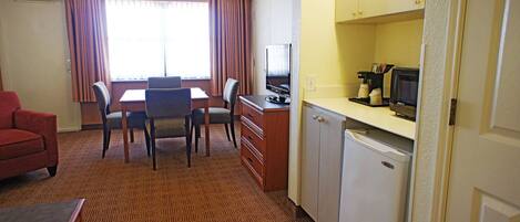 A hotel room with a dining table, chairs, a TV, a mini-fridge, and a red sofa.