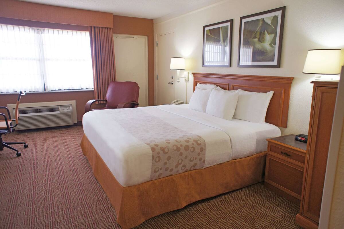 A hotel room with a large bed, a desk, a chair, a lamp, and a heater.