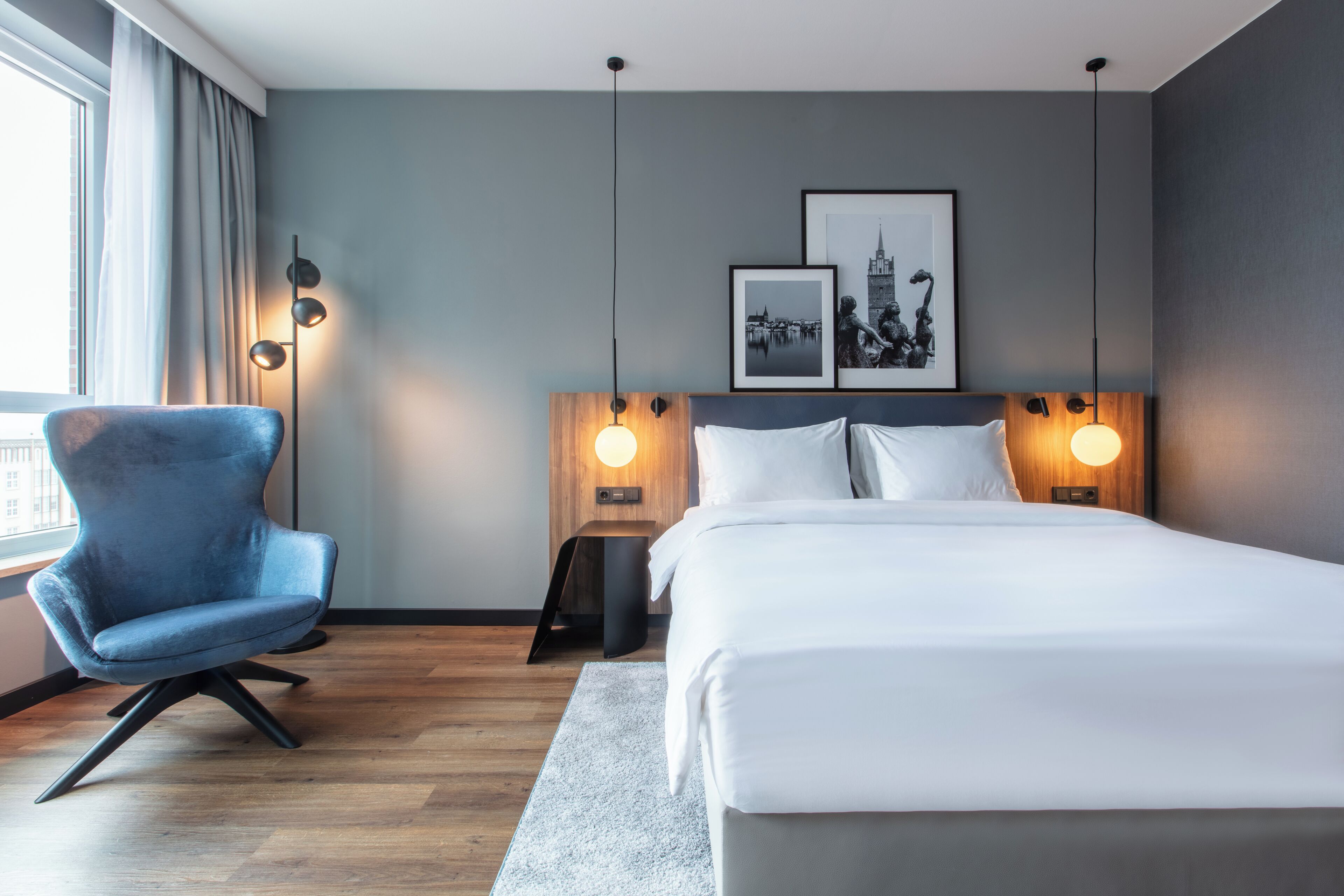 Una habitación de hotel moderna con una cama grande, un sillón azul y dos cuadros en la pared.