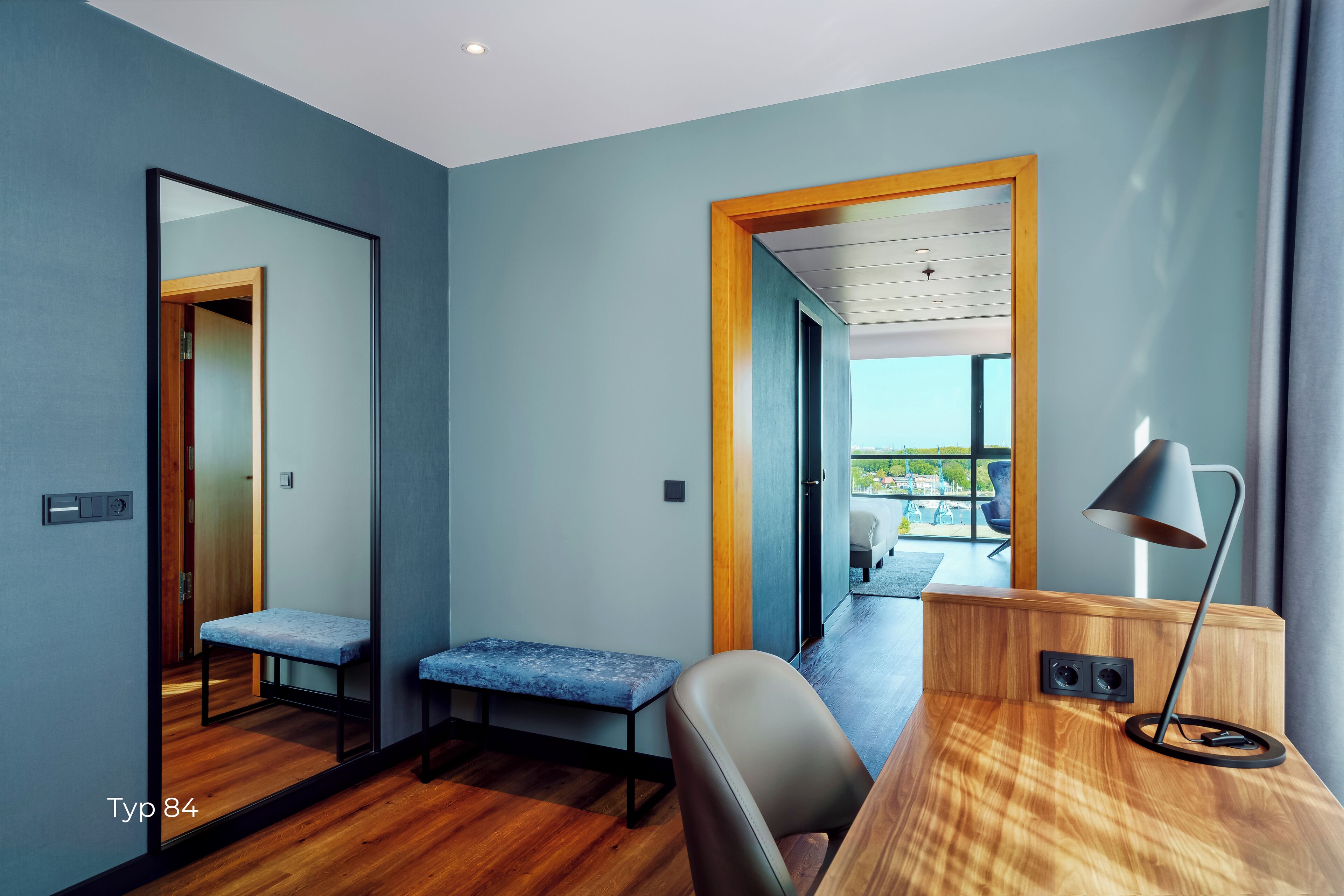A modern hotel room with a wooden desk, a bench, a mirror, and a view of the outdoors.