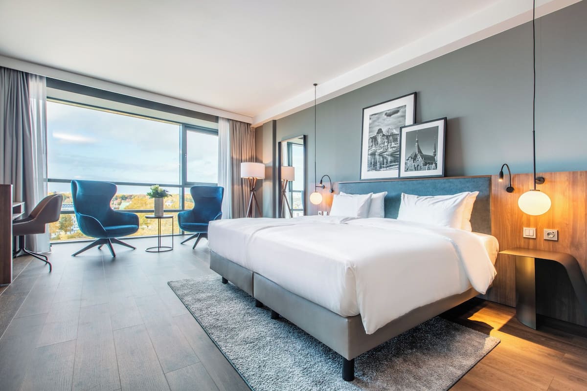 A modern hotel room with a large bed, two armchairs, a desk, and a view of the cityscape through large windows.