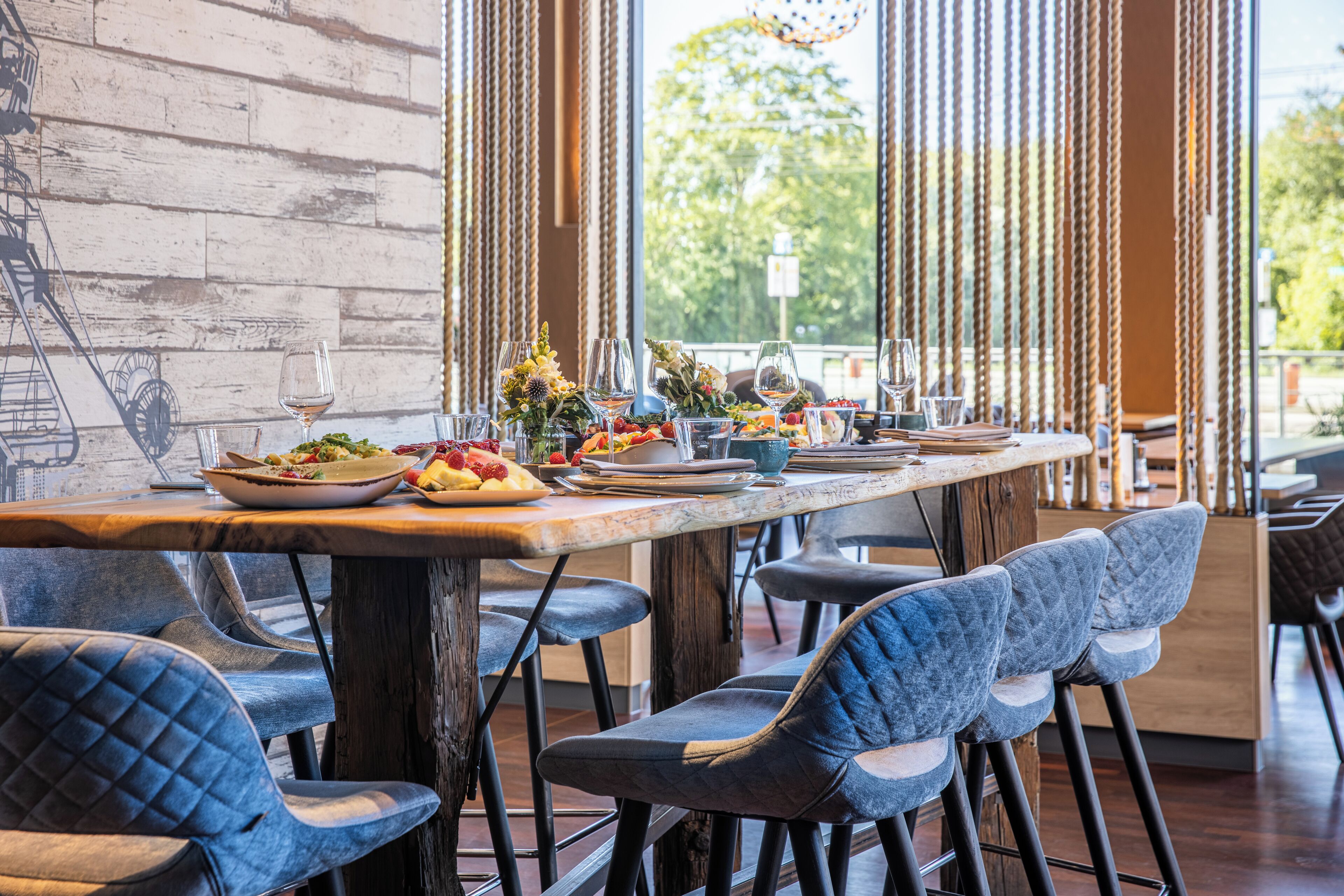 A restaurant with a long wooden table set for dining, blue upholstered chairs, and a view of greenery through large windows.