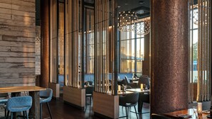 A modern restaurant with wooden tables and chairs, a brick column, and a large window with a view of the city. - ScanHotels City (Rostock)