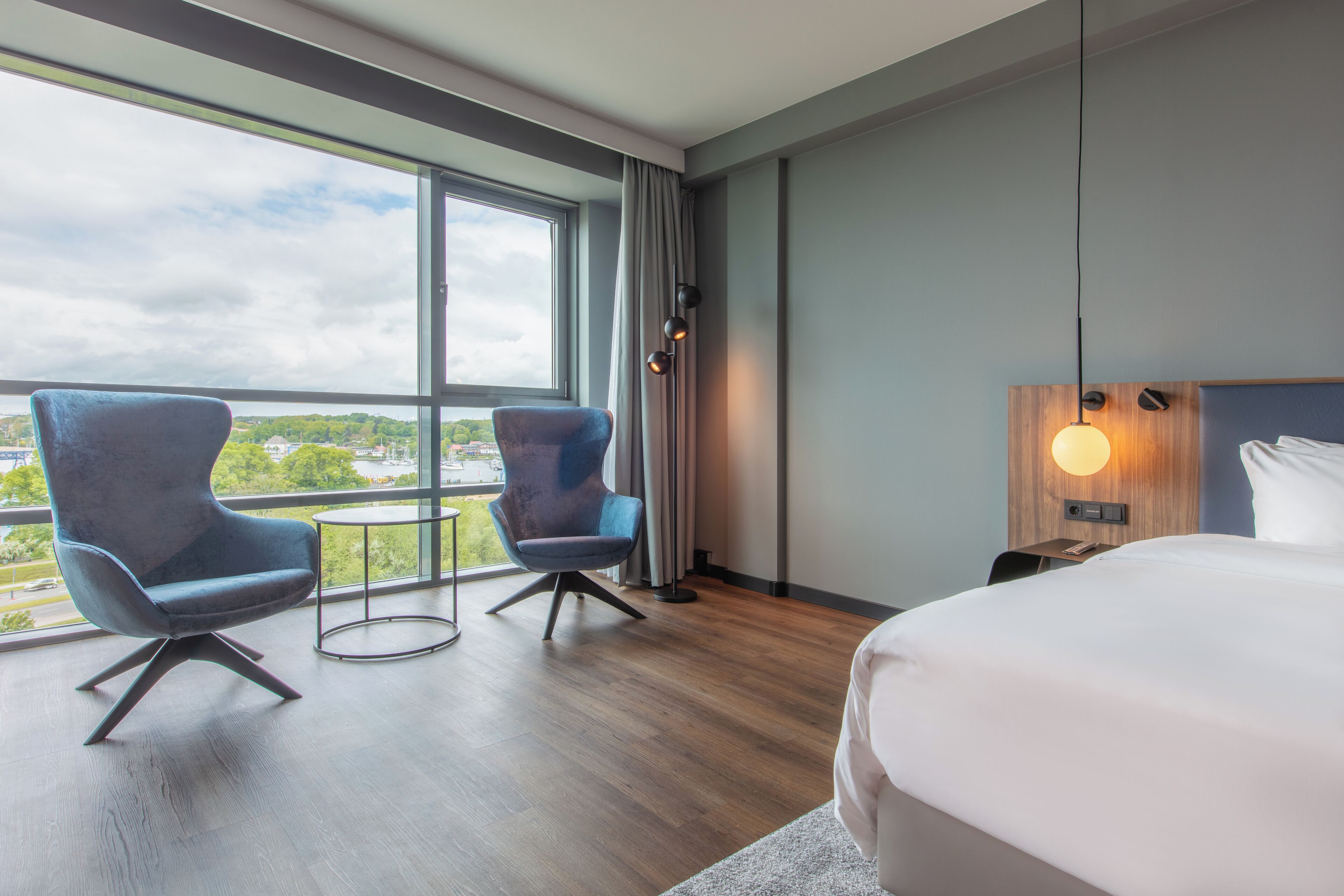 Una habitación de hotel moderna con una cama grande, dos sillones, una mesita y vistas a la ciudad a través de amplios ventanales.