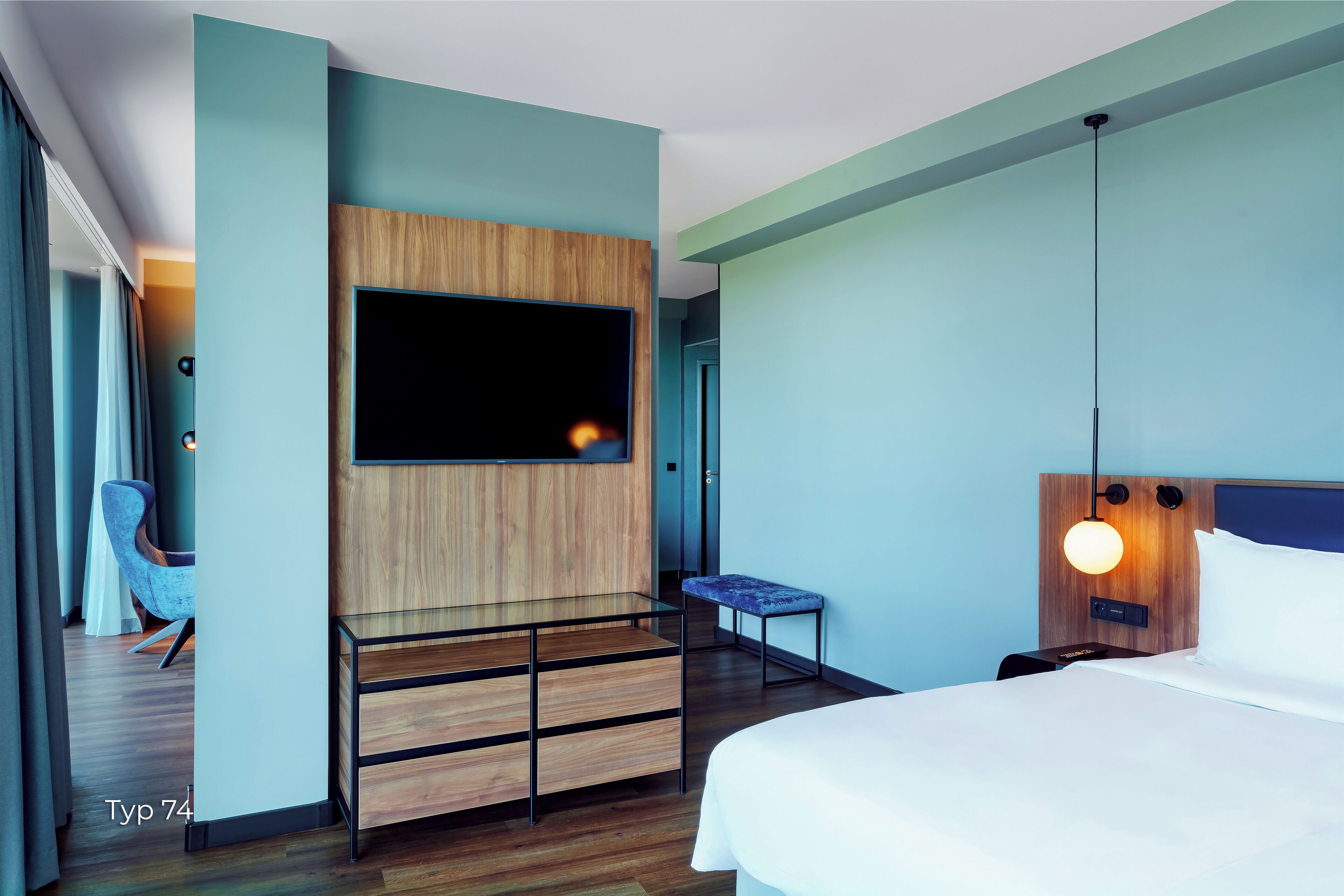 A modern hotel room with a large bed, a wooden TV stand, a small bench, and a wall-mounted light fixture.