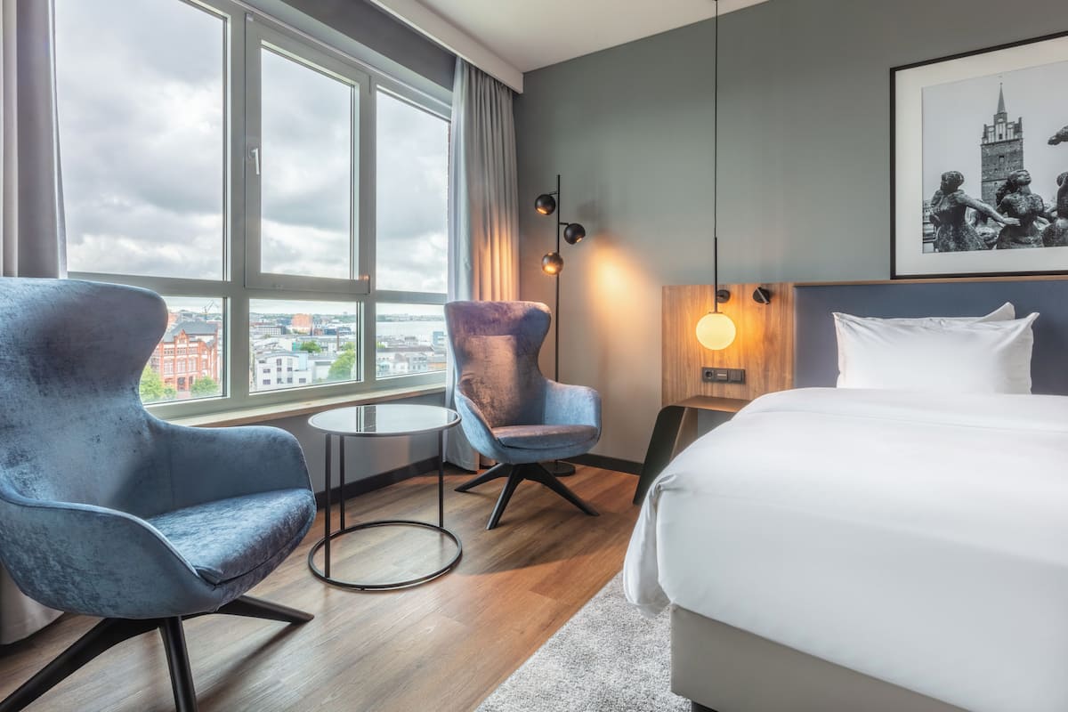A hotel room with a large window, a bed, a blue armchair, a small round table, and a wall-mounted light fixture.