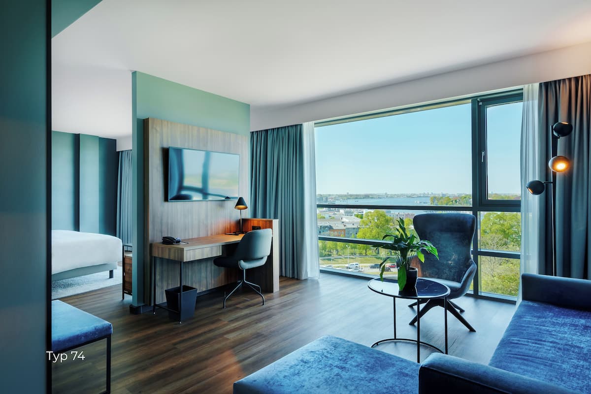 A modern hotel room with a bed, desk, chair, and a large window offering a city view.