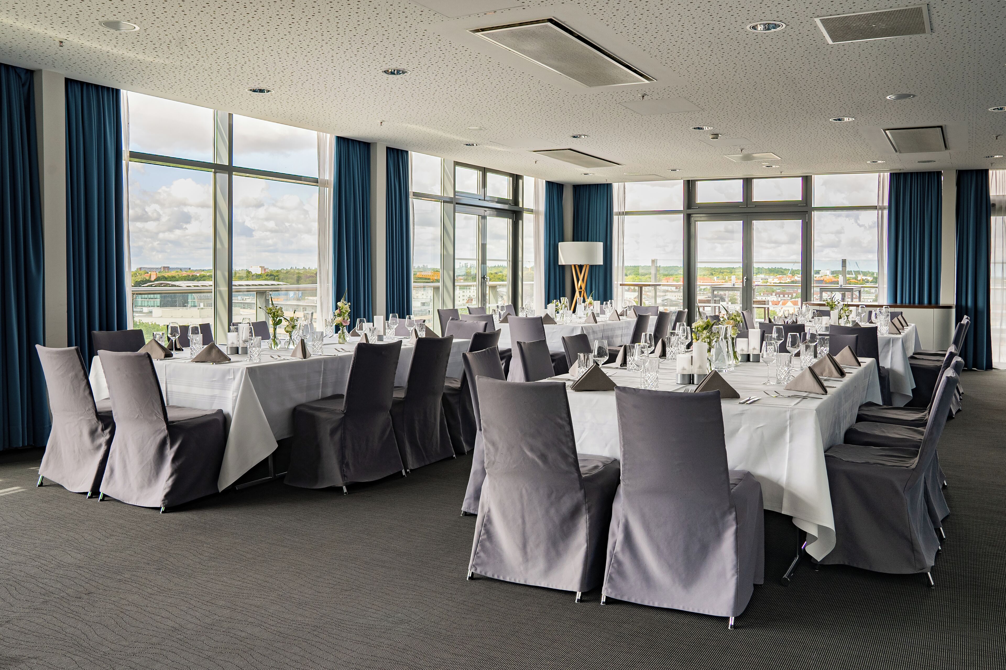 Un salón de conferencias con mesas dispuestas para una reunión, amplias ventanas con vistas y suelo alfombrado.