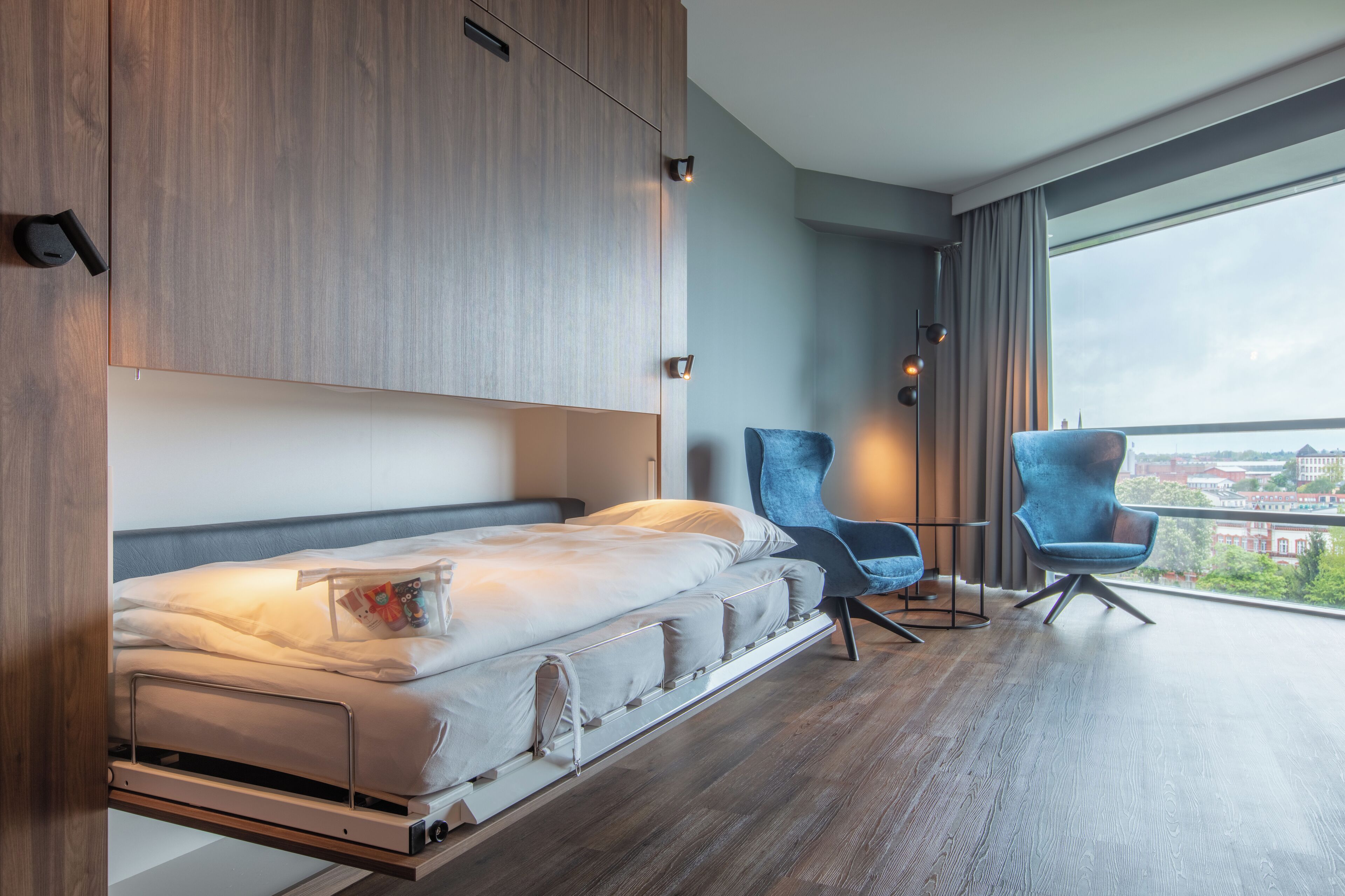 A modern bedroom with a bed, two chairs, and a large window offering a city view.