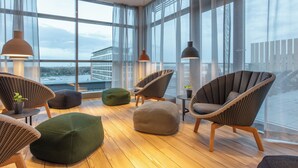 A modern lounge with wicker chairs, a green bean bag, and a view of a building across the water. - ScanHotels City (Rostock)