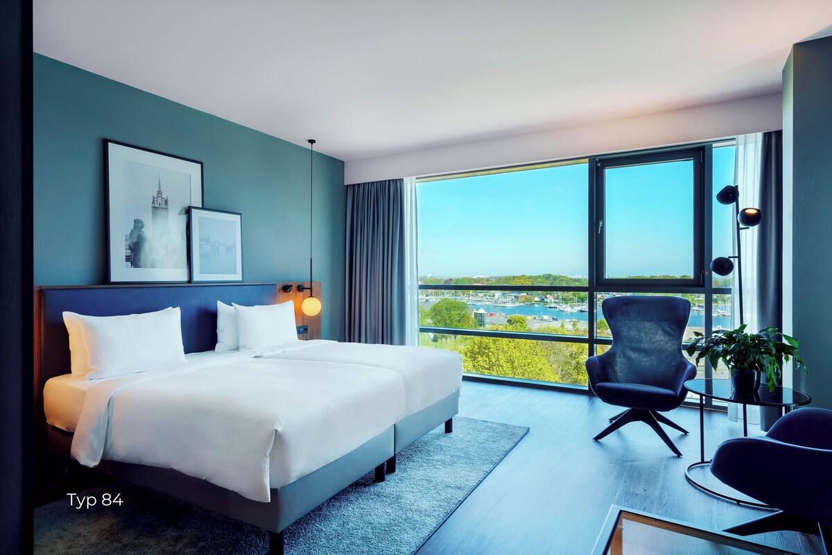 Ein modernes Hotelzimmer mit einem großen Bett, einem Sessel, einem kleinen Tisch und Blick auf die Stadt.