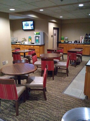 A hotel dining area with round tables and red chairs, a television on the wall, and a self-service beverage station.