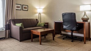A hotel room with a brown sofa, a wooden coffee table, a black office chair, a desk with a lamp, and two framed pictures on the wall. - Quality Inn Summerville - Charleston (Summerville)