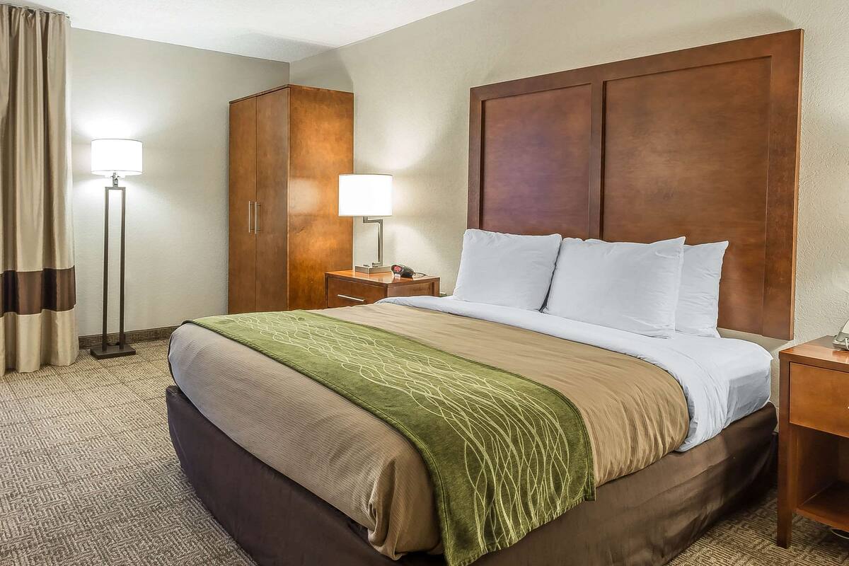A hotel room with a large bed, a wooden wardrobe, a nightstand with a lamp, and a floor lamp.