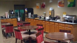 A hotel dining area with multiple tables and chairs, a kitchen counter with appliances, and various food and beverage dispensers. - Quality Inn Summerville - Charleston (Summerville)
