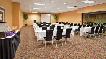 A conference room with rows of tables and chairs, a projector screen, and a plant.