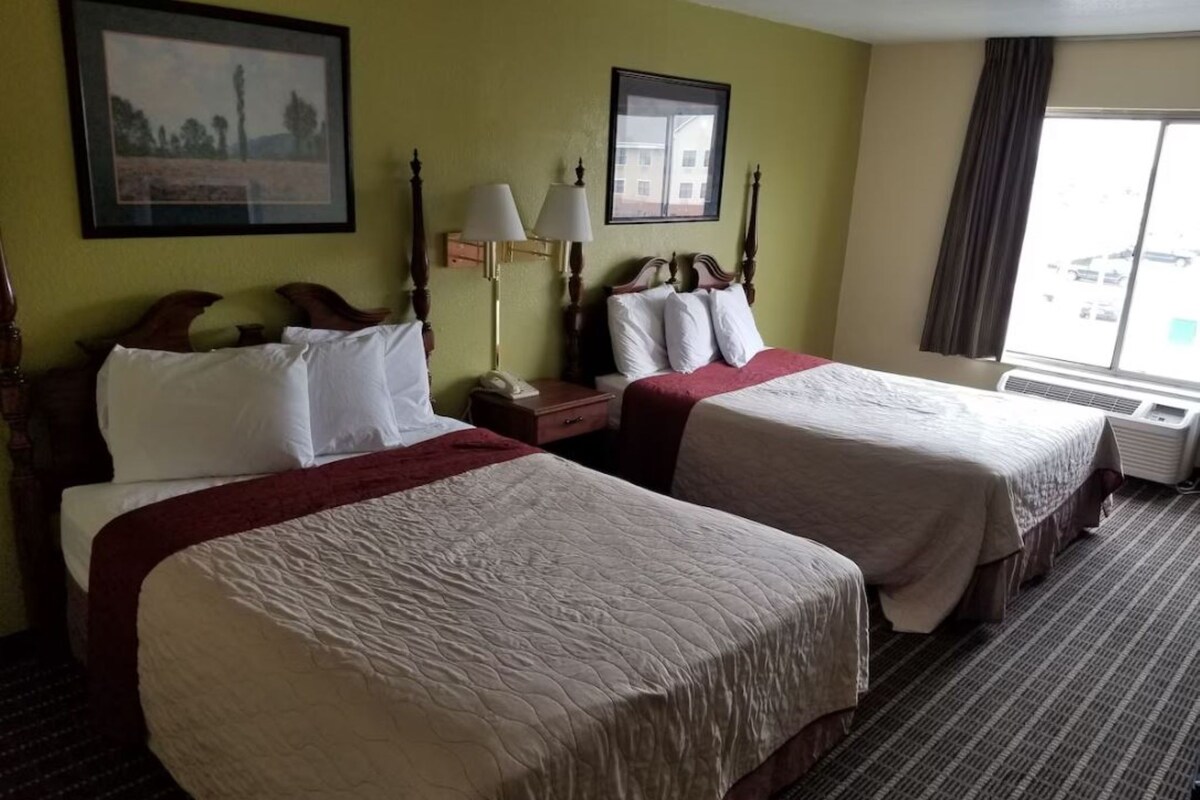 A hotel room with two beds, a nightstand, a lamp, and a window with a view.