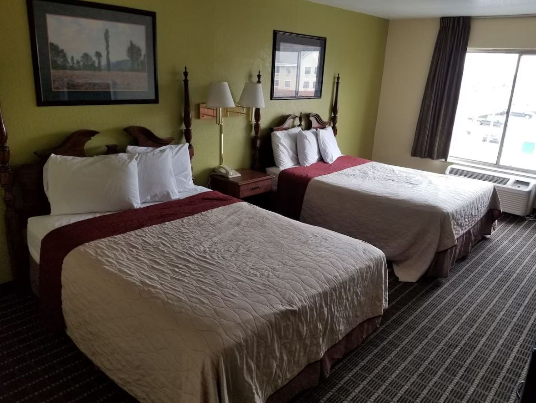 A hotel room with two beds, a nightstand, a lamp, and a window with a view.
