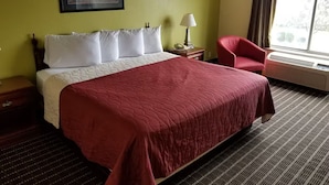 A hotel room with a bed, a red chair, a nightstand, a lamp, and a window with curtains.