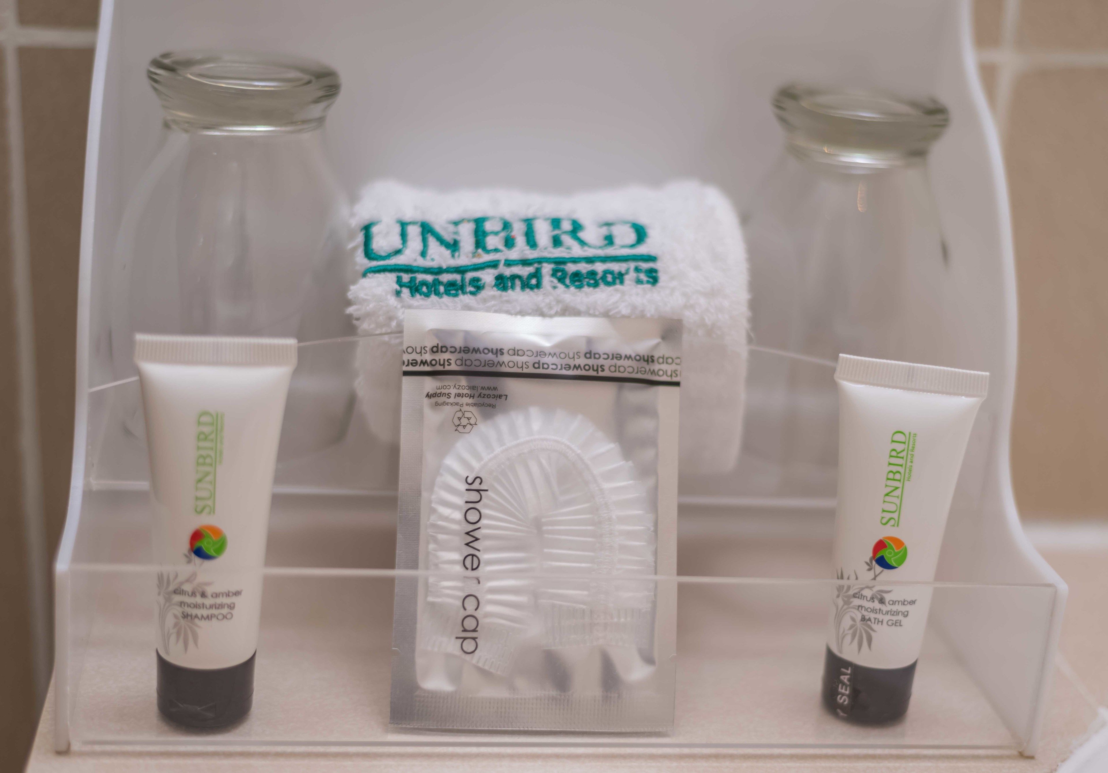 Un tocador de baño con un portalámparas de vidrio transparente que contiene champú y gel de baño hidratante Sunbird, un gorro de ducha blanco y una toalla con el logo de Sunbird.