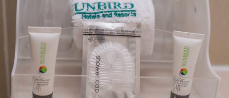 Un tocador de baño con un portalámparas de vidrio transparente que contiene champú y gel de baño hidratante Sunbird, un gorro de ducha blanco y una toalla con el logo de Sunbird.