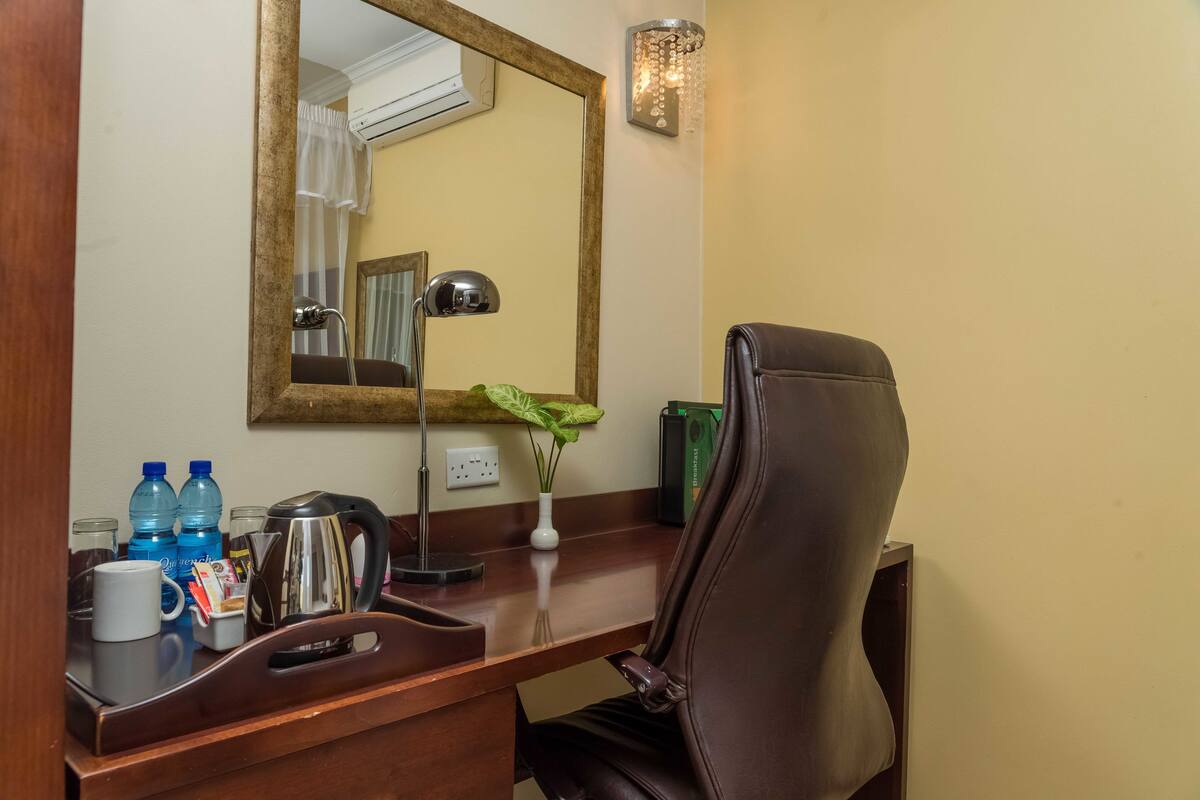 Escritorio de habitación de hotel con espejo, lámpara, silla, florero con planta y artículos de aseo.