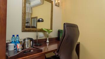 A hotel room desk with a mirror, a lamp, a chair, a vase with a plant, and some toiletries.