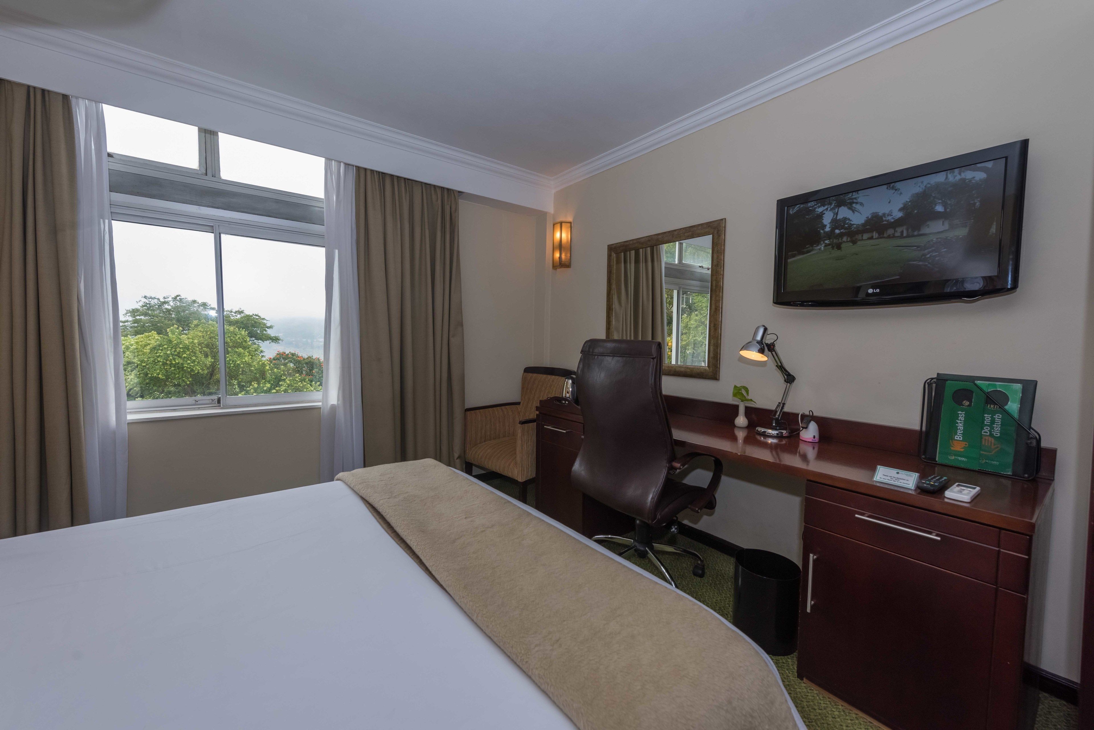 A hotel room with a bed, desk, chair, TV, and a view of greenery outside.