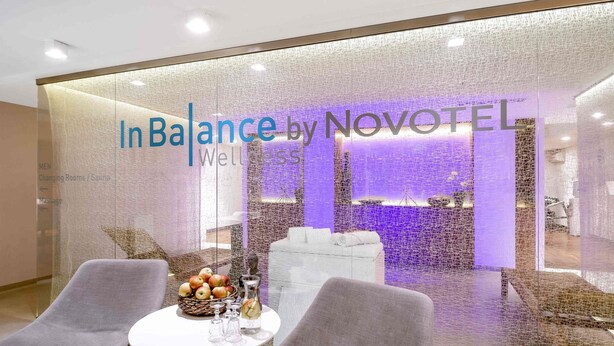 A modern interior with a glass wall displaying 'In Balance by NOVOTEL Wellness.' There are two chairs, a small table with a fruit basket, and a Buddha statue.
