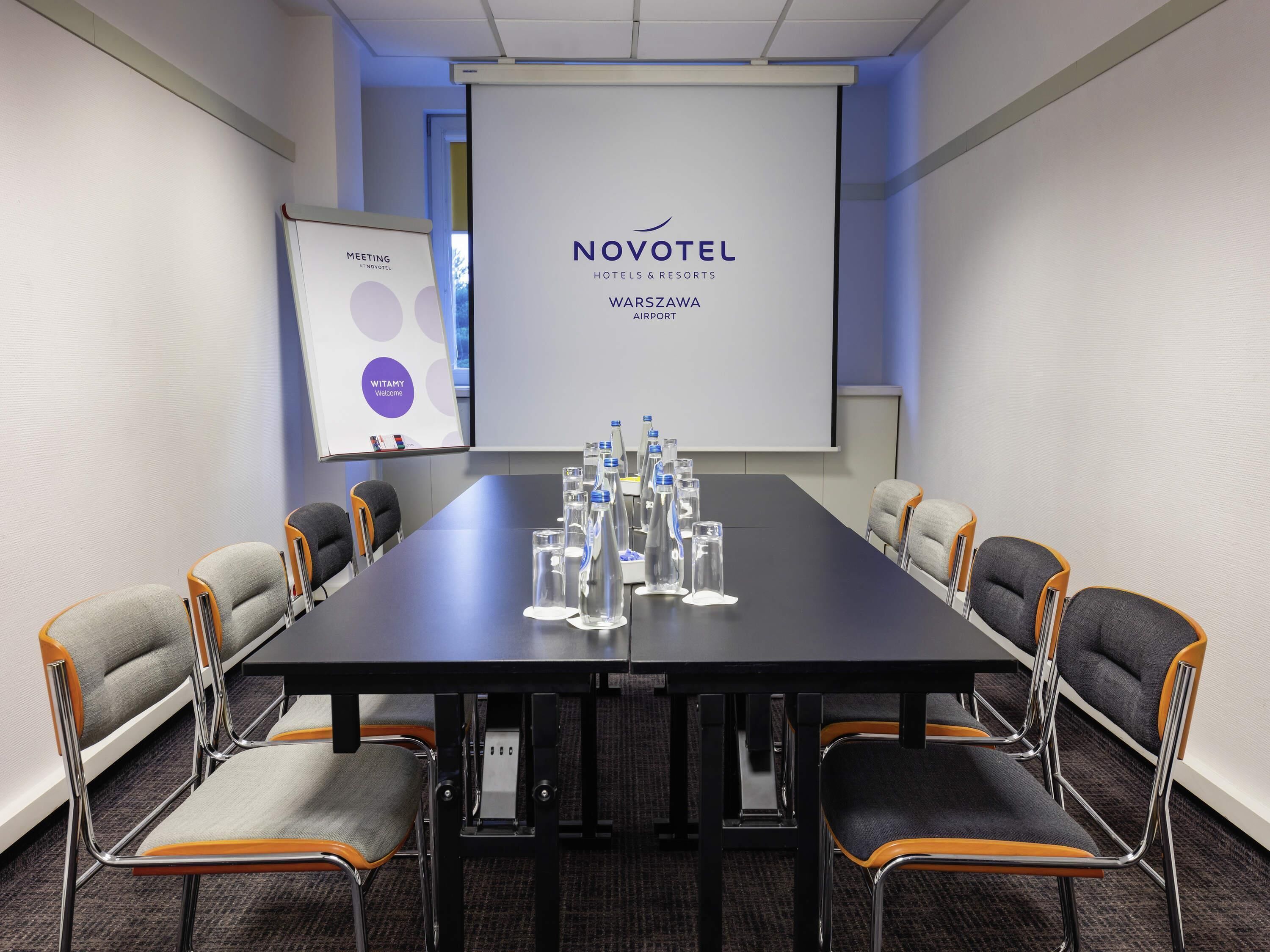 A meeting room with a Novotel logo, a projector screen, a whiteboard, and water bottles on the table.