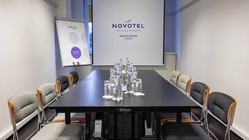 A meeting room with a Novotel logo, a projector screen, a whiteboard, and water bottles on the table.
