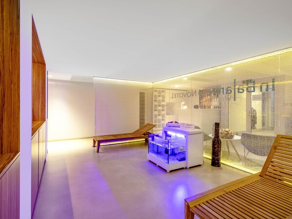 A modern spa room with a wooden bench, a massage table, and a glass enclosure with a water feature.