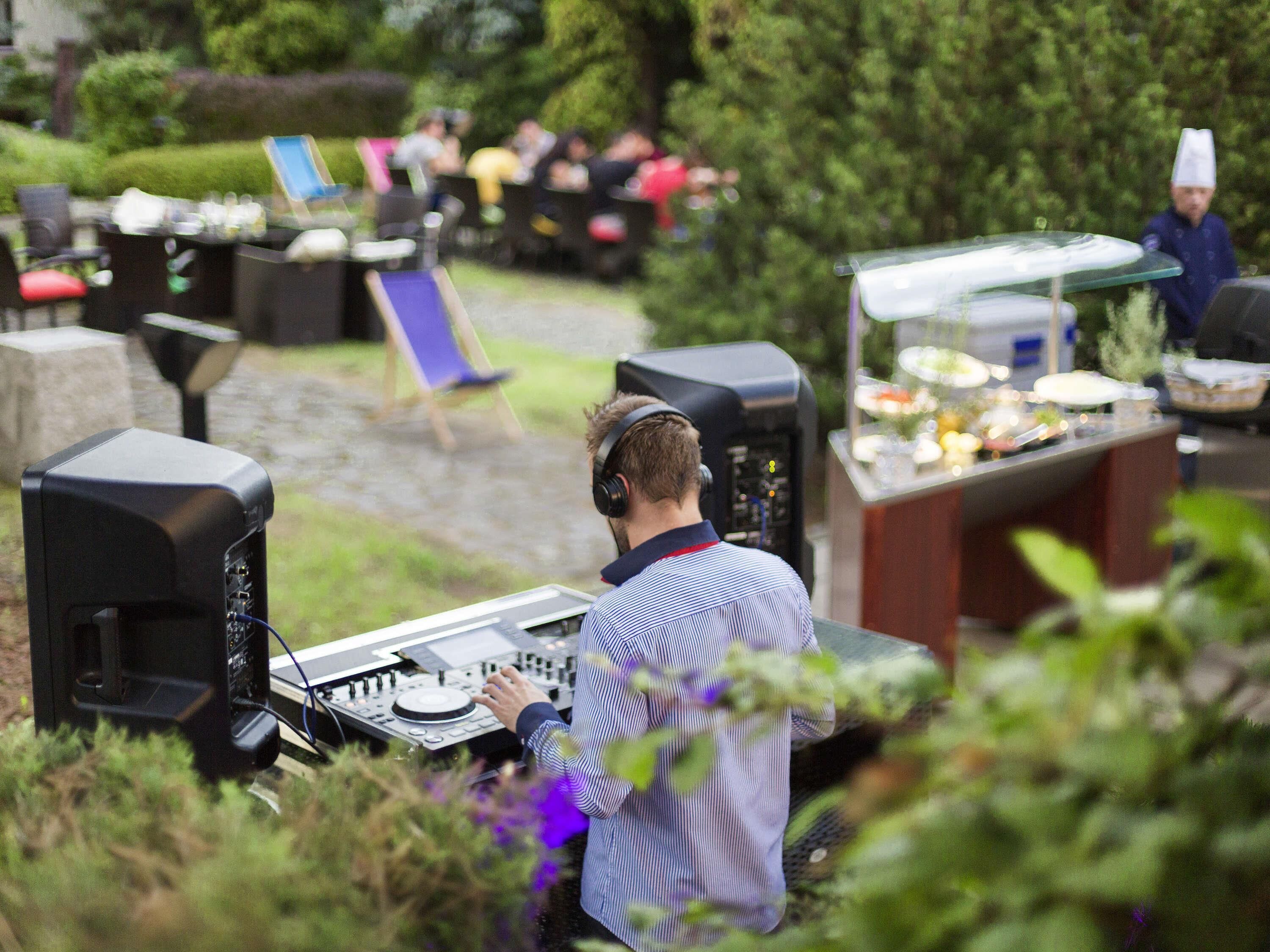A DJ booth with audio equipment and a laptop, surrounded by outdoor seating and a food service area.
