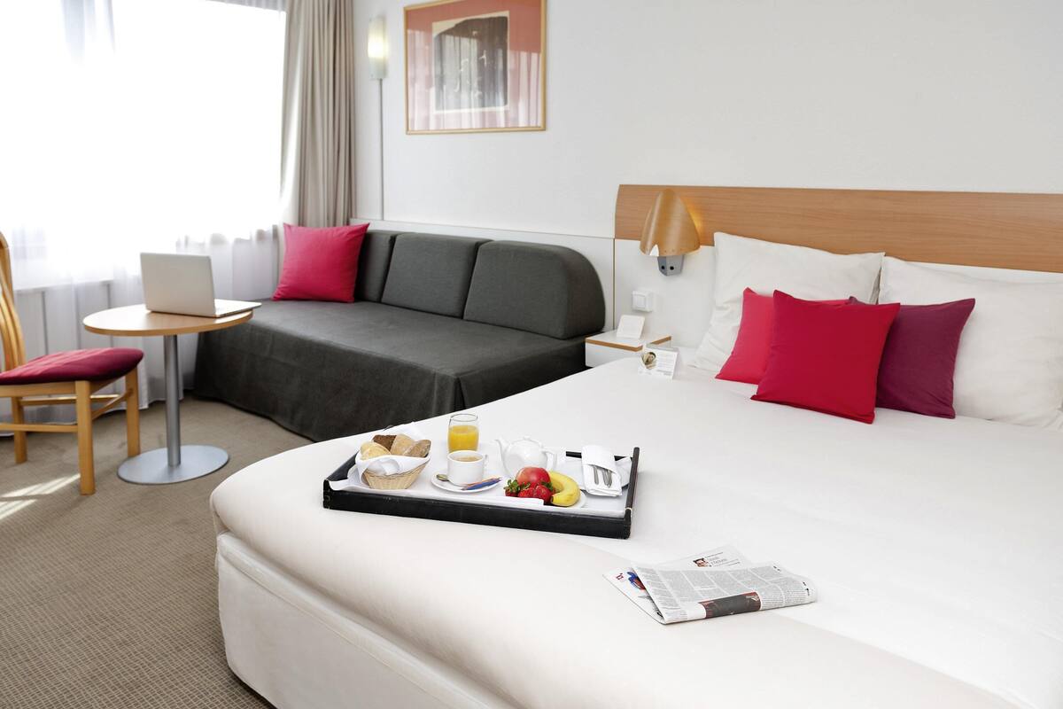 Ett hotellrum med en säng, en soffa, ett skrivbord med en bärbar dator, ett brickbord med frukost och en sänglampa.