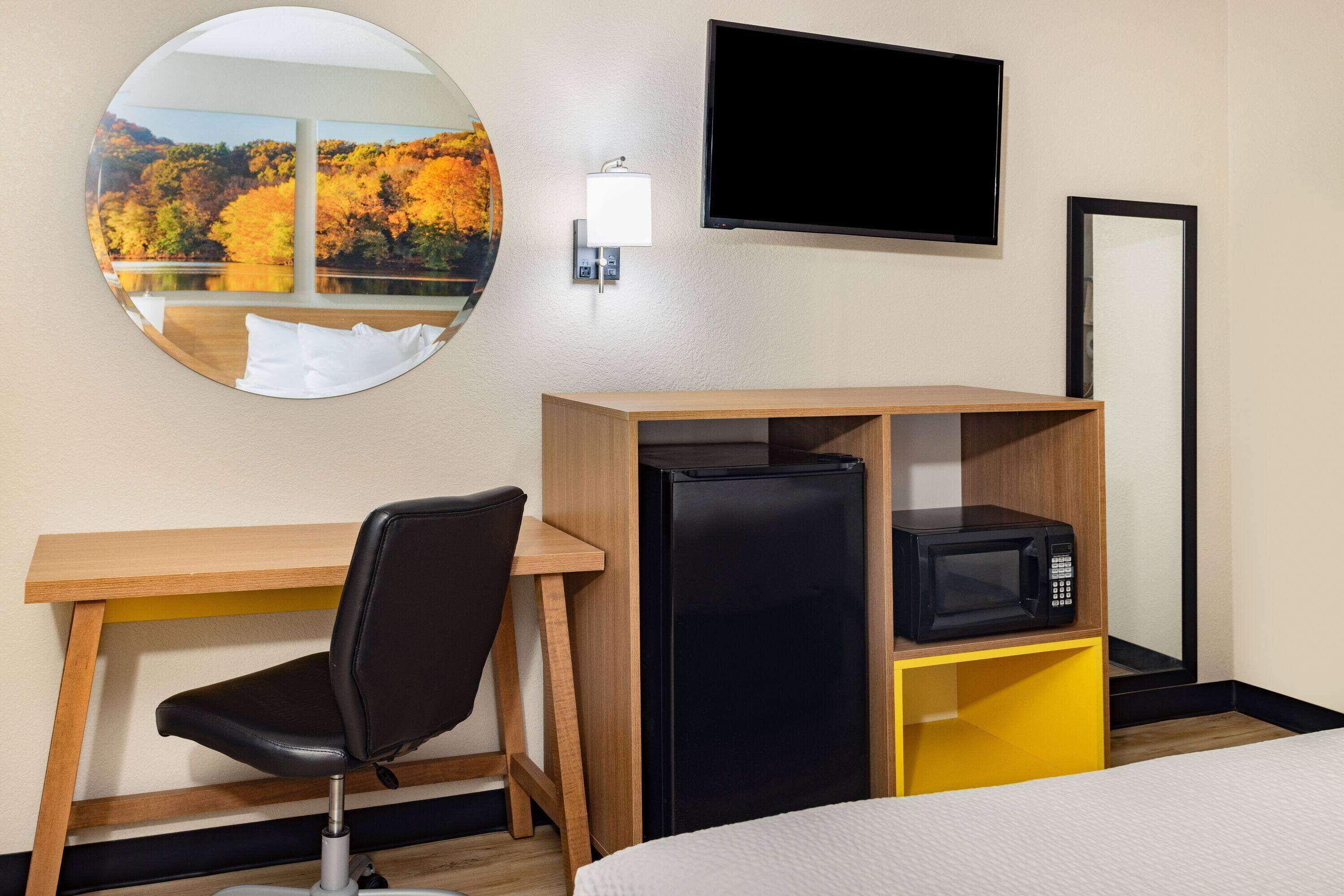 Quarto de hotel com cama, escrivaninha com cadeira, frigorífico preto, micro-ondas, televisão e espelho redondo com vista panorâmica.