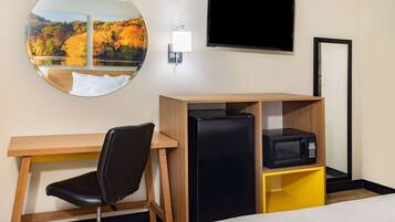 A hotel room with a bed, a desk with a chair, a black refrigerator, a microwave, a TV, and a round mirror with a scenic view.