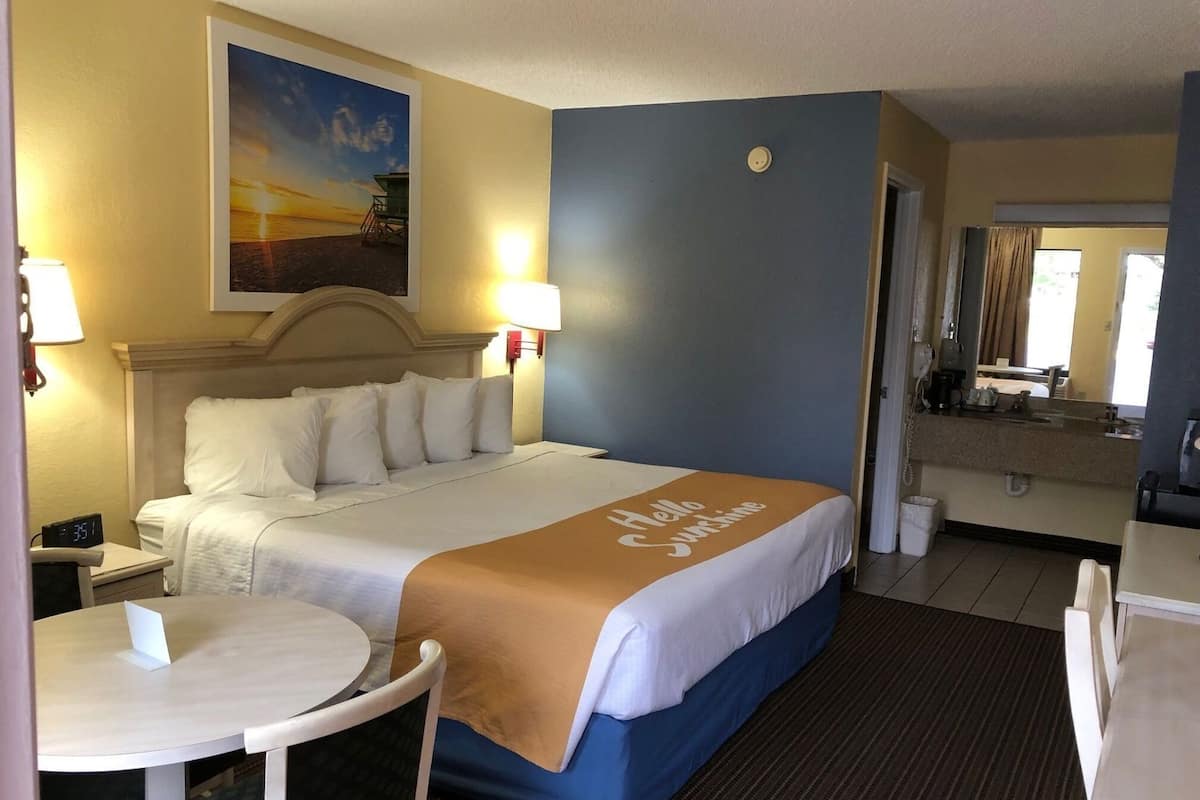 Habitación de hotel con cama, una mesa redonda, mesita de noche, lámpara, un cuadro, un espejo y un baño visible a través de una puerta abierta.