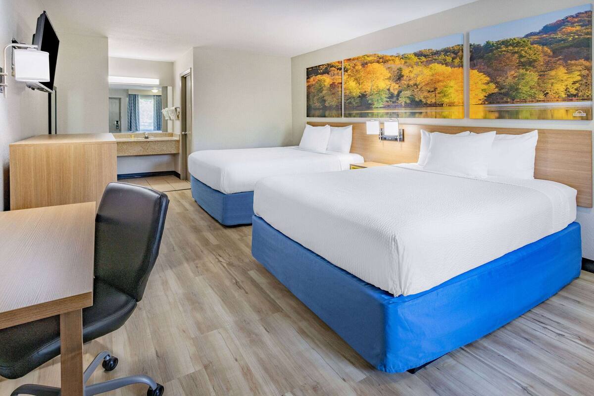 A hotel room with two beds, a desk, a chair, and a large window with autumn foliage artwork.