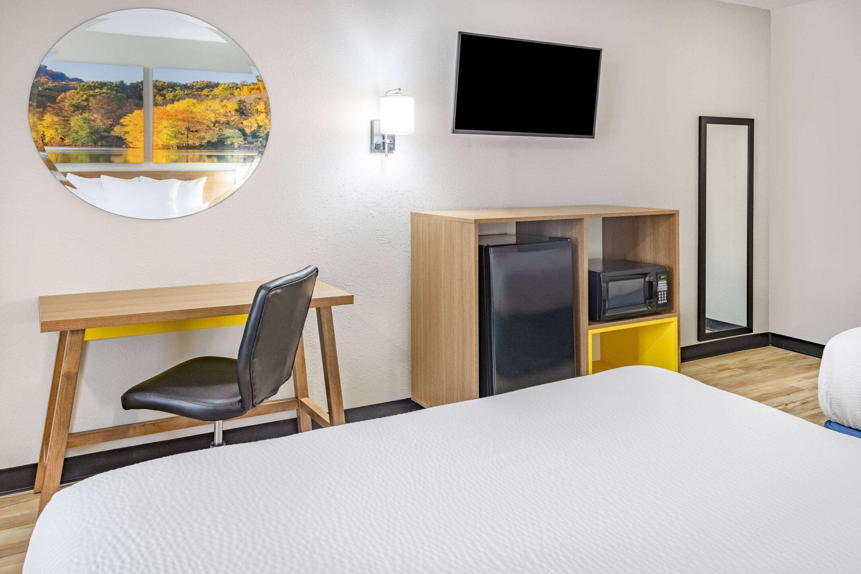A hotel room with a bed, a desk with a chair, a TV mounted on the wall, a microwave, and a refrigerator.