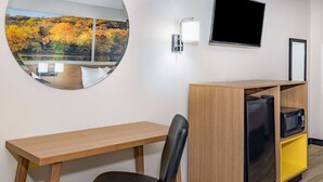 Habitación de hotel con escritorio de madera, una silla negra, un refrigerador pequeño, microondas y televisión en la pared.